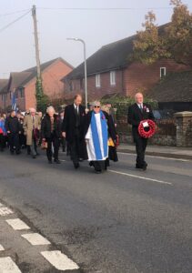 People walking along road in parade with man at front holding red poppy wreath