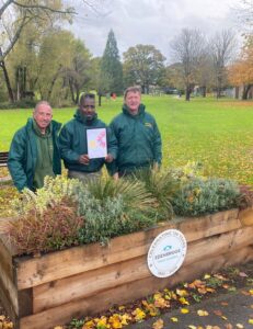 grounds team men wearing green work jackets in front of planter with shrubs holding certificate