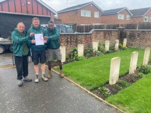 grounds team men wearing green work jackets in front of cemetery holding certificate