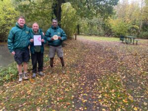 grounds team men wearing green work jackets in front of park with pond holding certificate