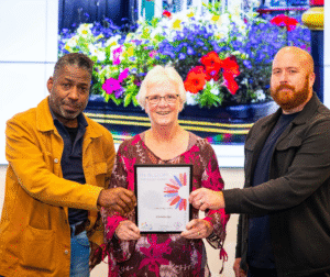 man with yellow jacket, lady and flower blouse and man in black jacket holding certificate with photo of flower planter in background