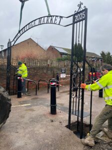 Two men in high-vis jackets putting iron archway with words Edenbridge Market in ground
