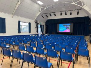 Community hall with blue chairs lined up and presentation screen on stage