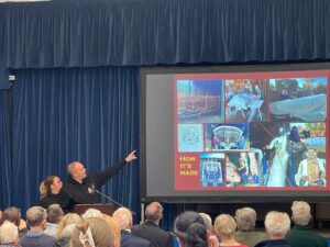Lady and man pointing to a presentation screen in front of audience sat on chairs