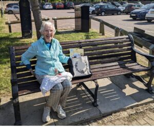 older lady sitting on bench in car park smiling with shopping bag