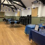 Interior of hall with wooden floor, tables and chairs and bunting. Wine glasses on blue tablecloth