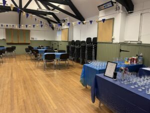 Interior of hall with wooden floor, tables and chairs and bunting. Wine glasses on blue tablecloth