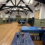 Interior of hall with wooden floor, tables and chairs and bunting. Wine glasses on blue tablecloth