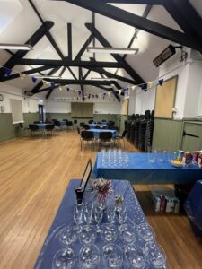Interior of hall with wooden floor, tables and chairs and bunting. Wine glasses on blue tablecloth