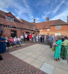 Lady in blue dress talking into microphone with group of people outside brick building