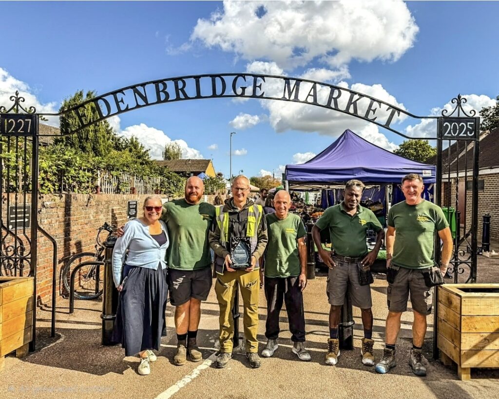 Six people standing in front of iron archway sign that says Edenbridge Market. The middle person is holding a glass trophy award