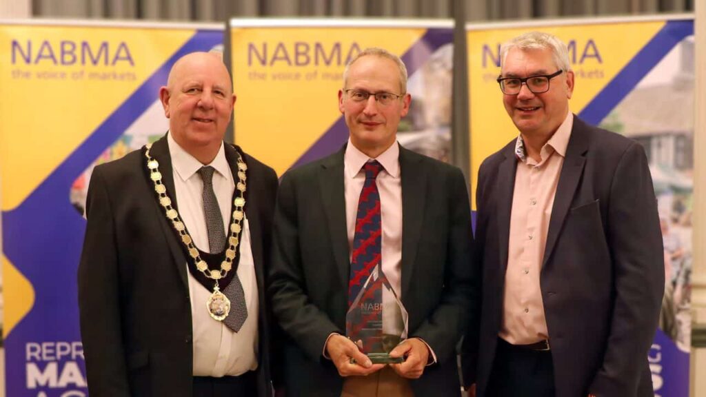 Three men in suits standing in front of awards banner. Man in middle holding a glass trophy that says Market Team of the Year